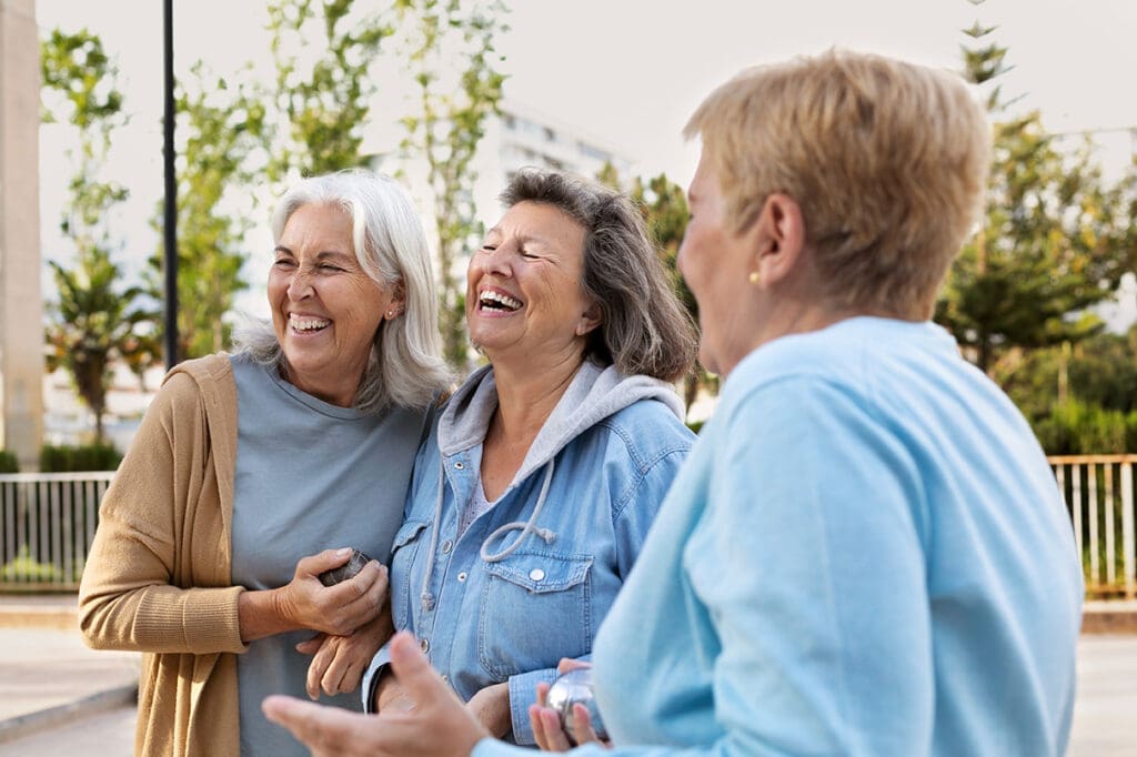 Three older women laughing together in a sunny park, enjoying each other's company and the outdoors.