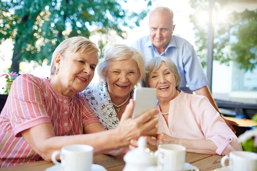 Three older women smiling and posing together while taking a selfie with a smartphone.
